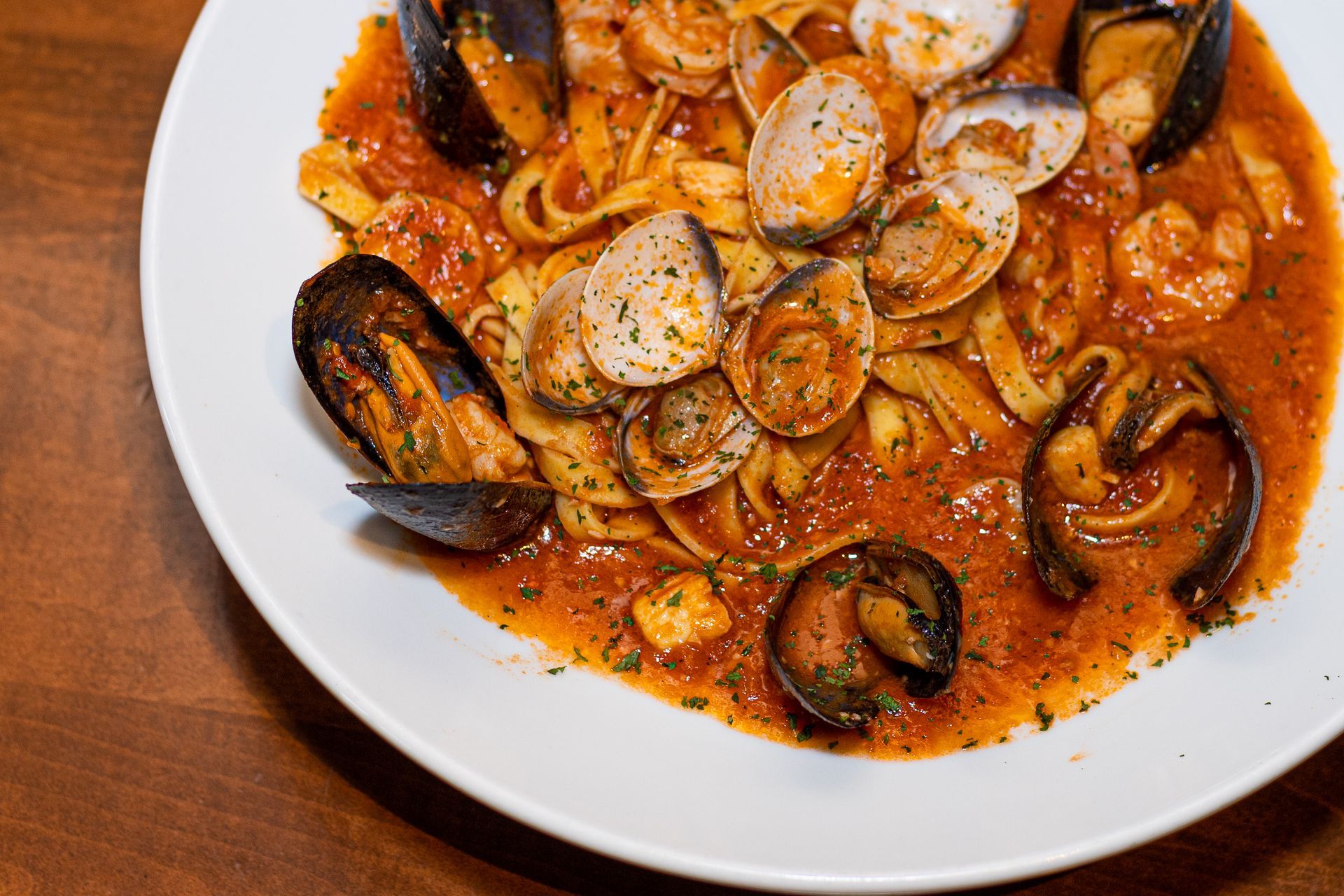 A white plate topped with noodles , mussels , shrimp and sauce on a wooden table.