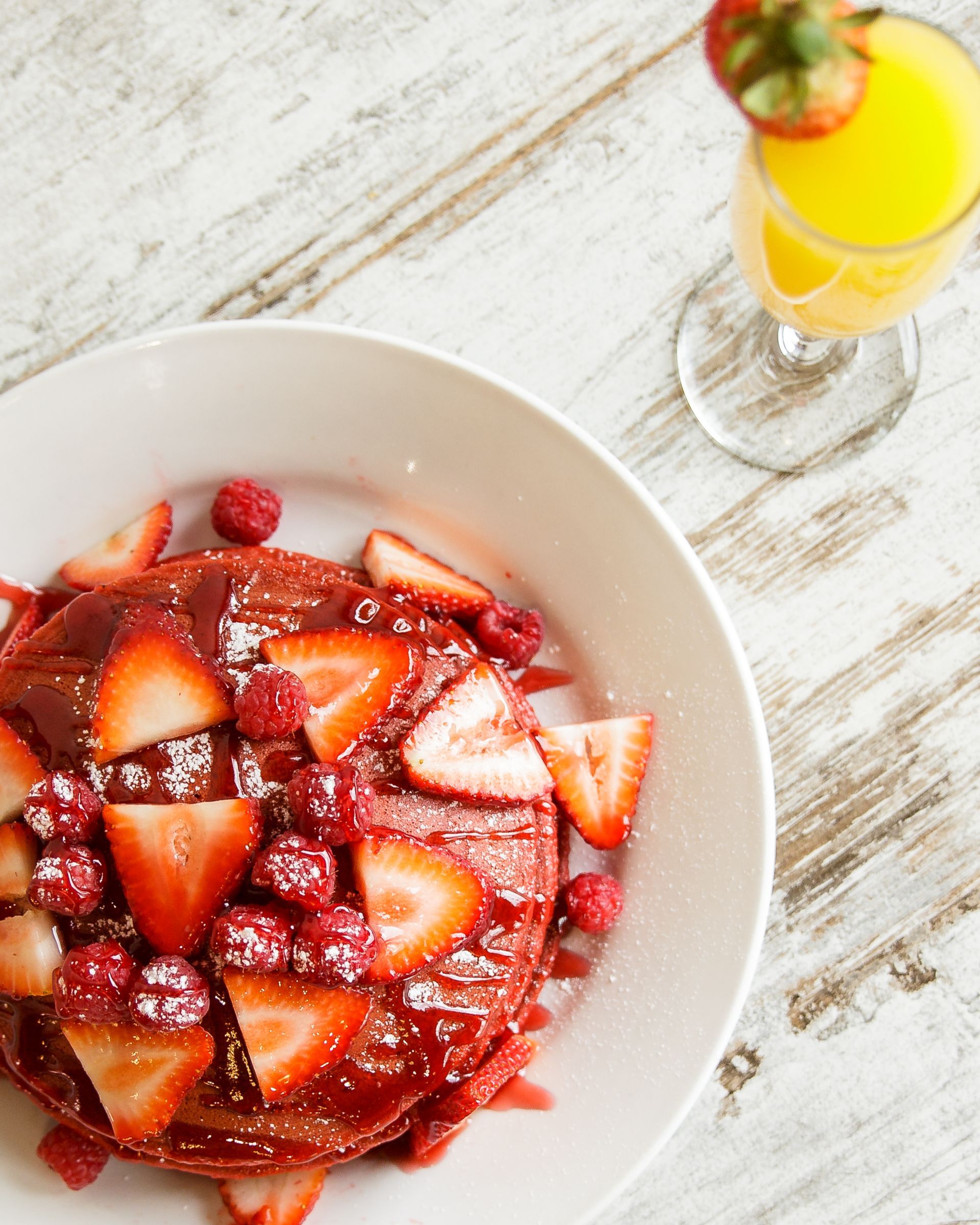 A plate of pancakes with strawberries and raspberries next to a glass of orange juice