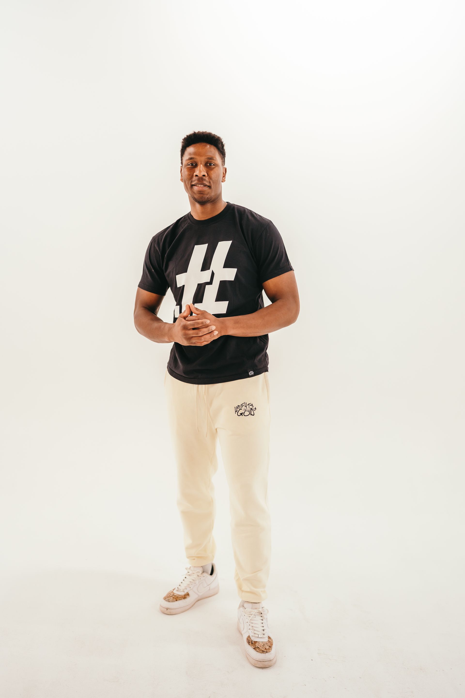 A man wearing a black t-shirt and white pants is standing in front of a white wall.