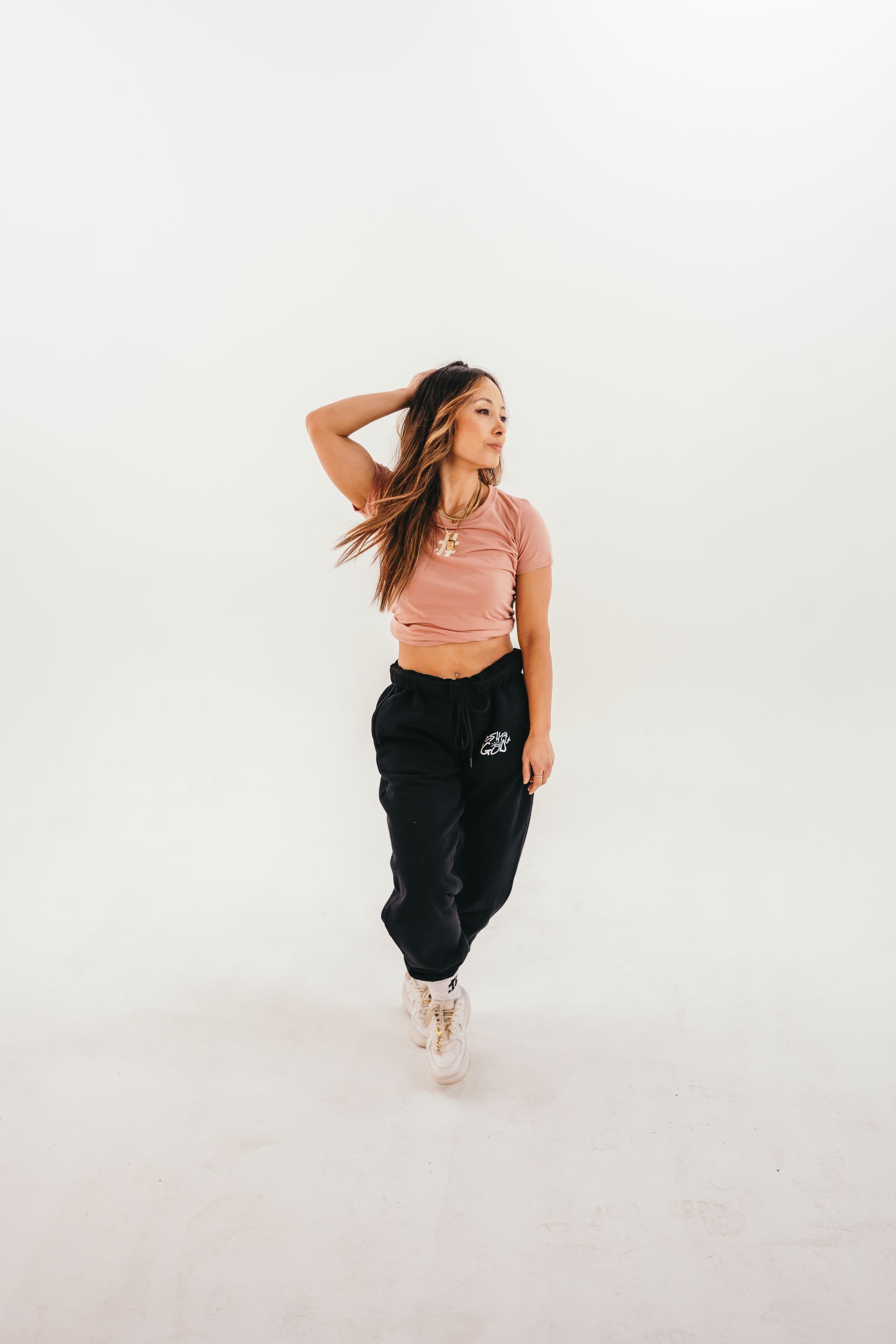 A woman in a pink crop top and black sweatpants is standing in front of a white wall.