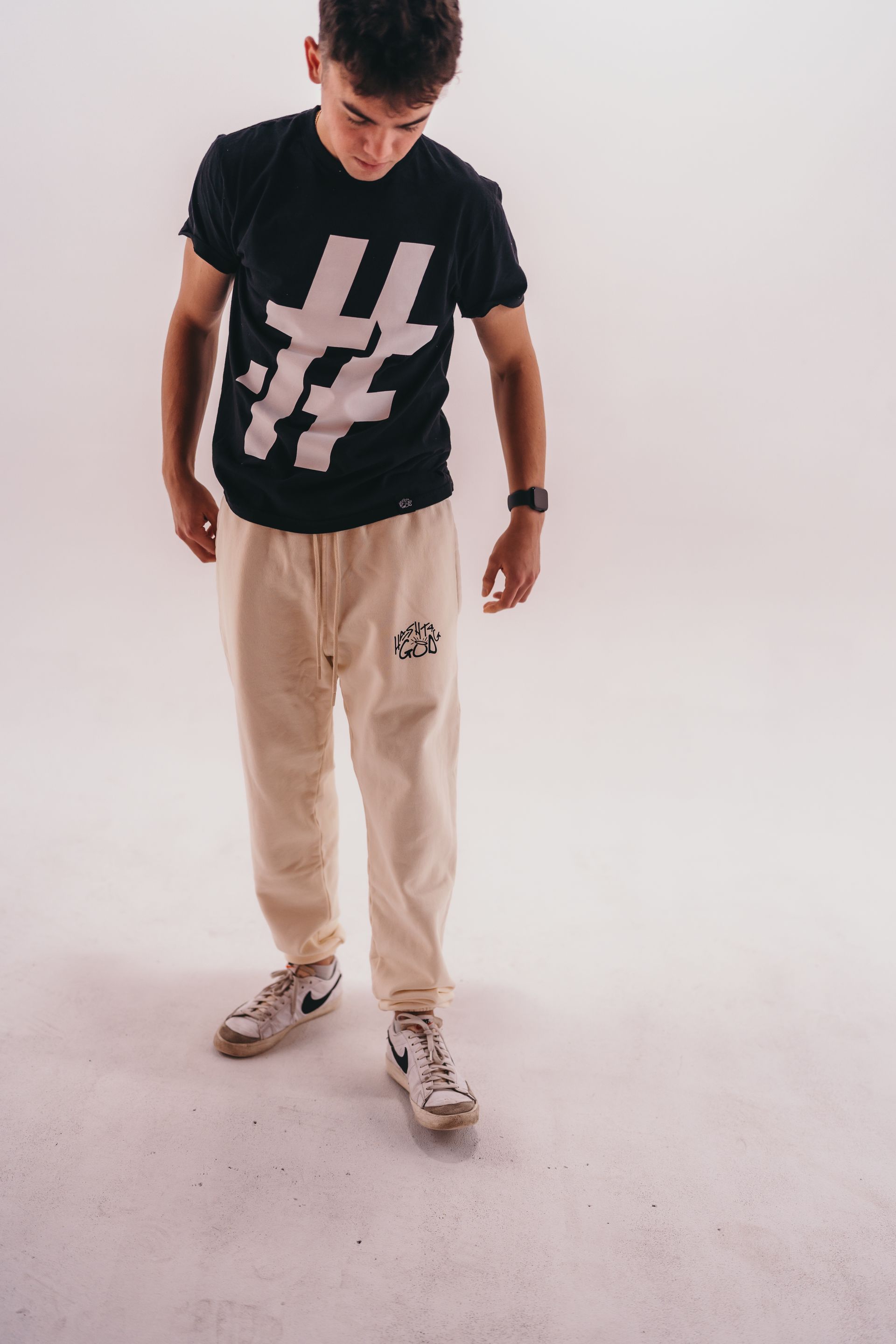 A young man wearing a black t-shirt and beige sweatpants is standing on a white surface.