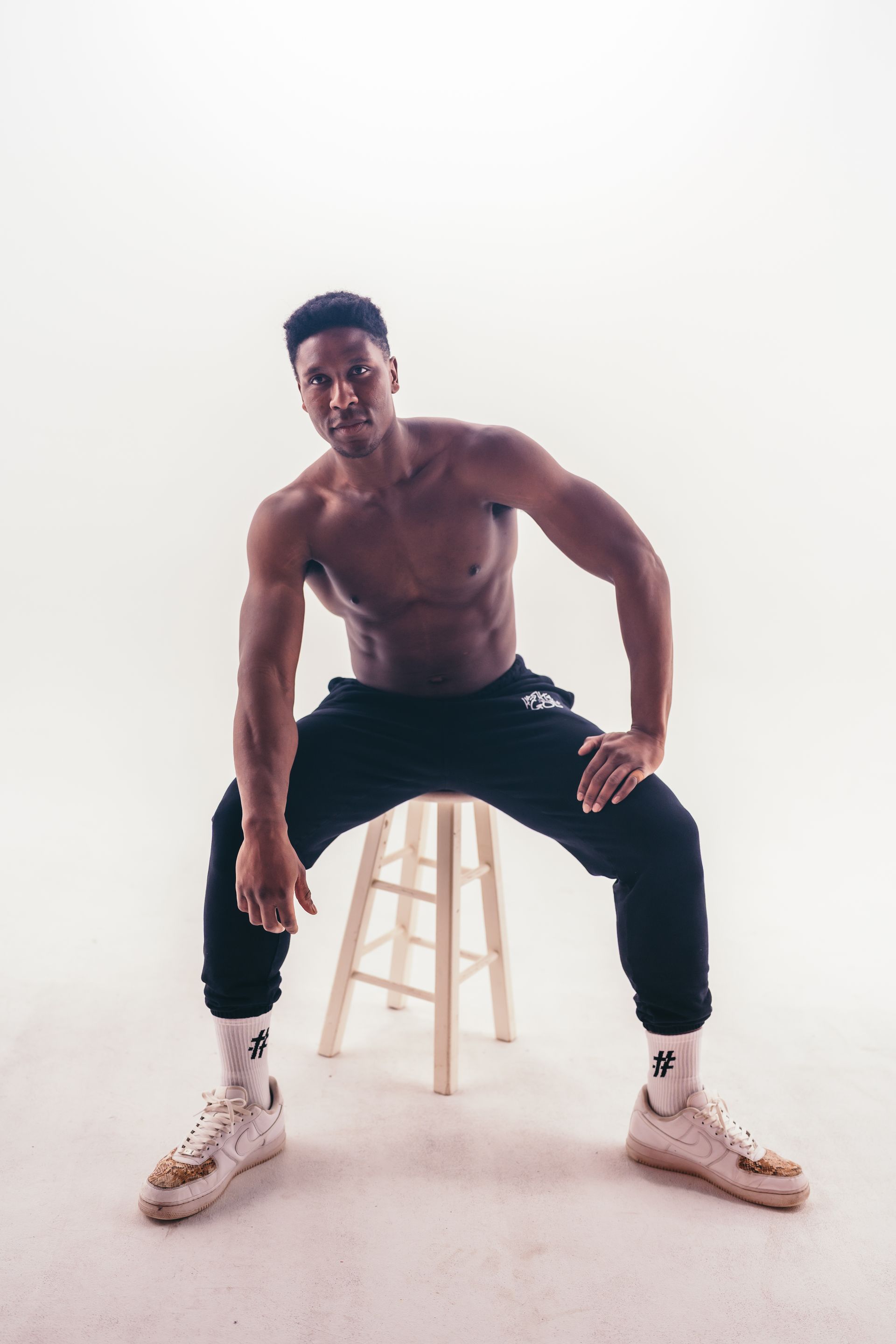 A shirtless man is sitting on a wooden stool.