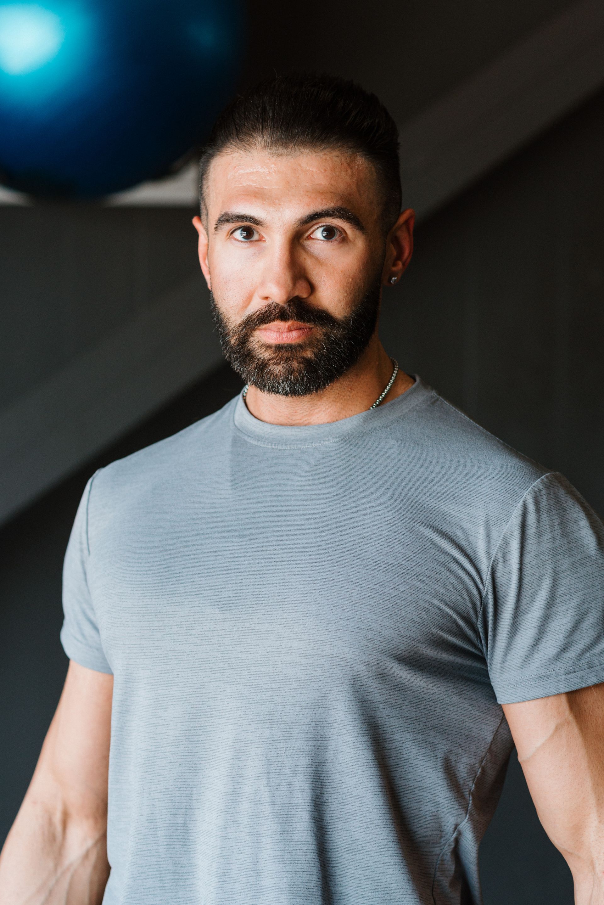 A man with a beard is wearing a grey t-shirt and standing in front of a pilates ball.
