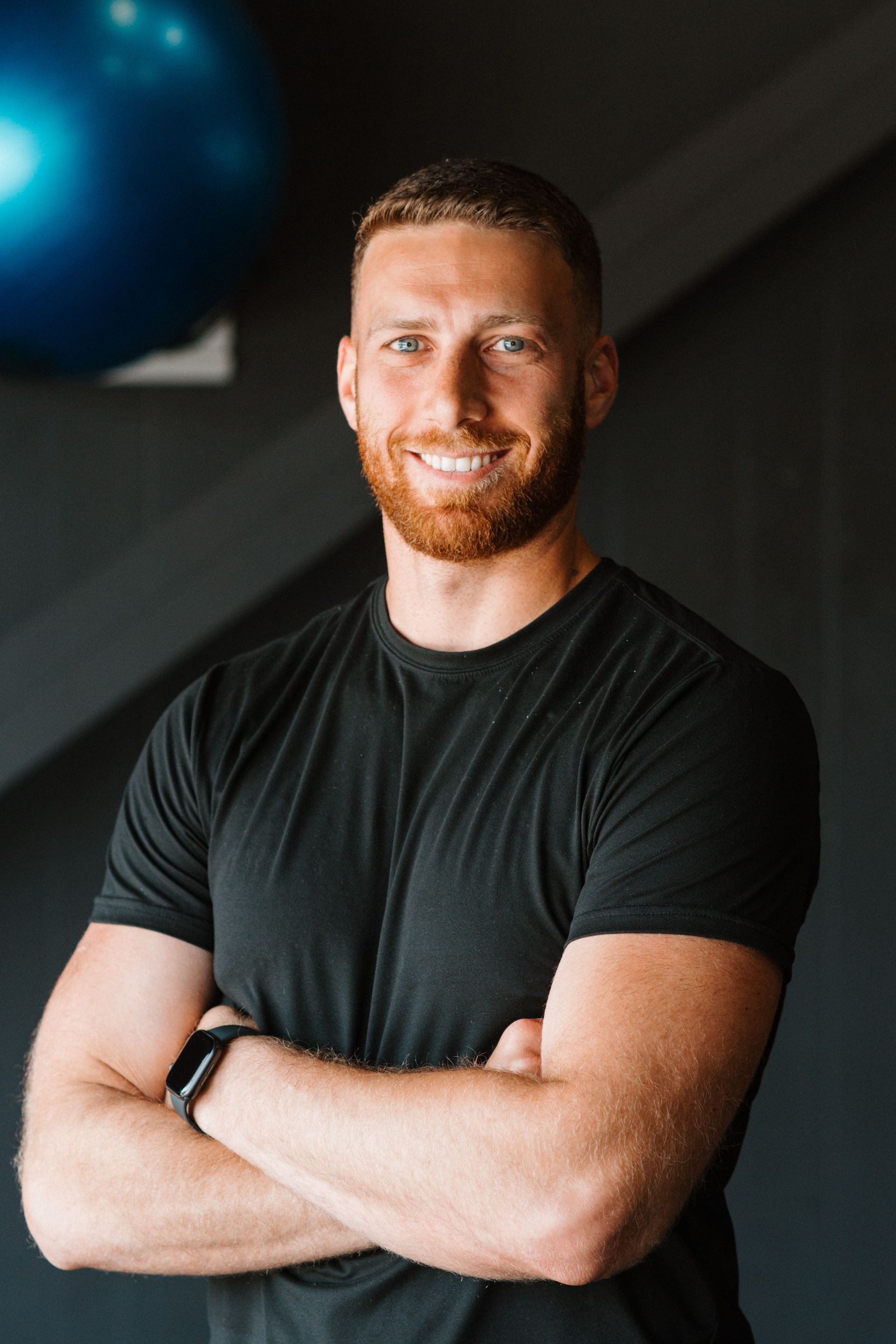 A man with a beard is standing with his arms crossed in a gym.