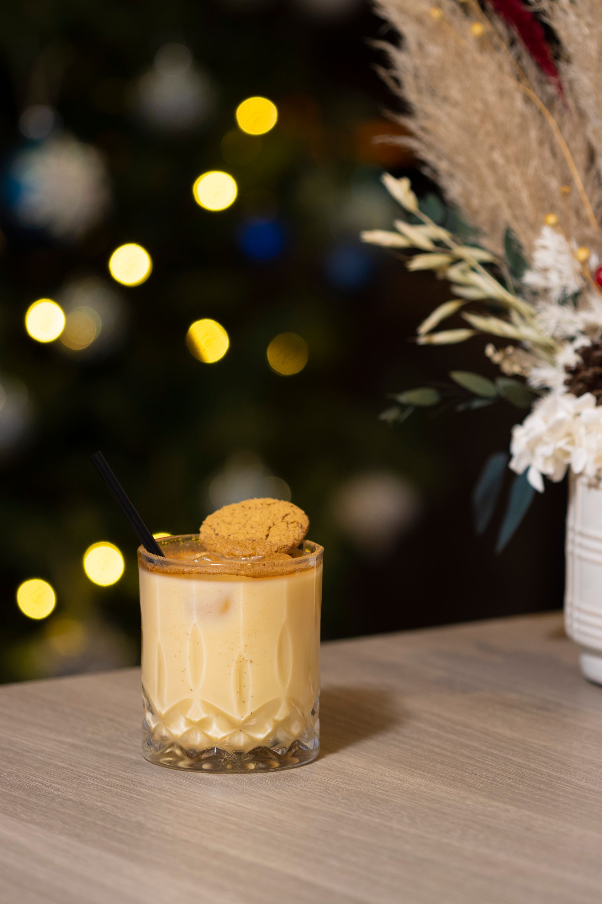 A glass of eggnog with a cookie on top is on a table in front of a christmas tree.