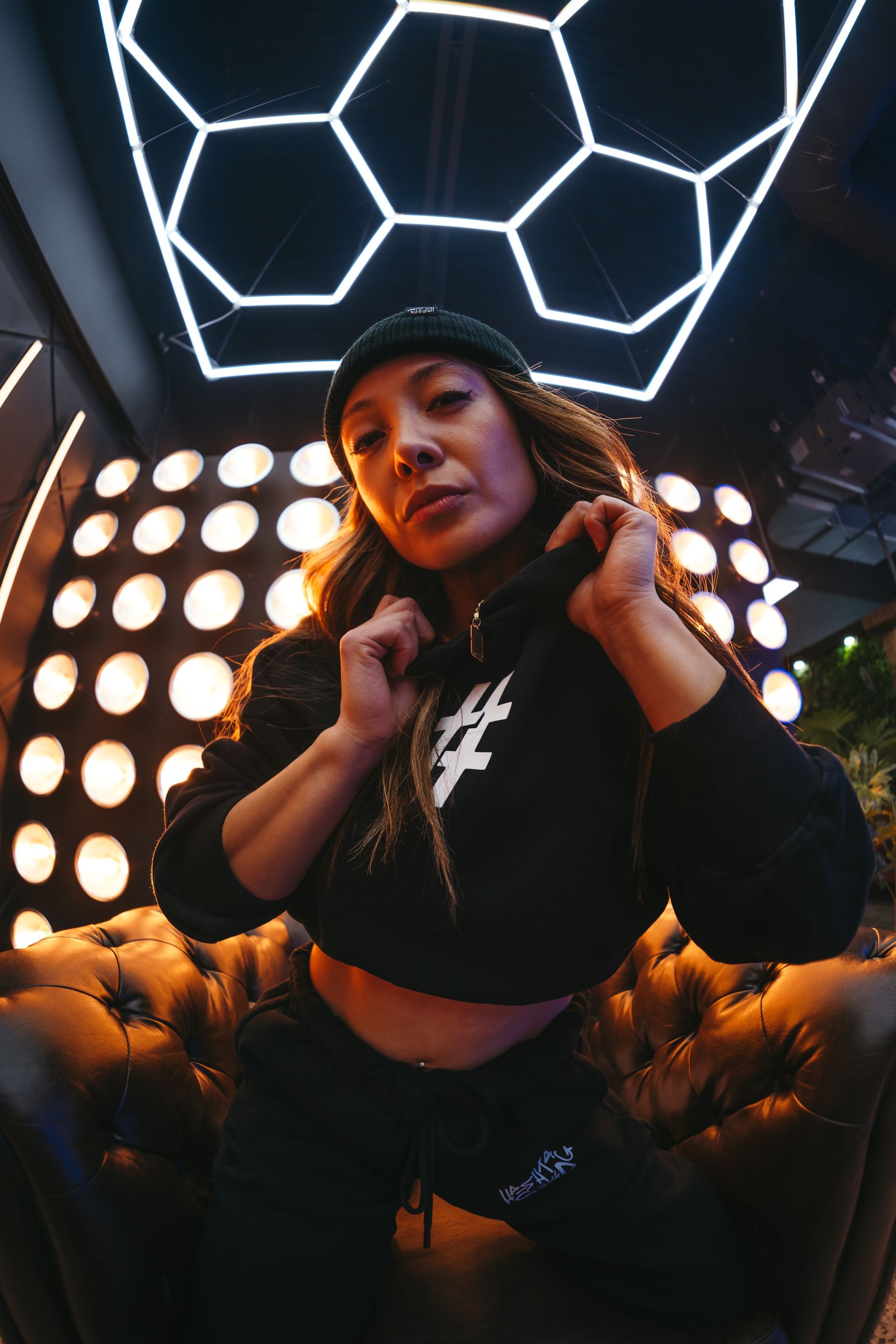 A woman is sitting on a couch wearing a black hoodie and a hat.