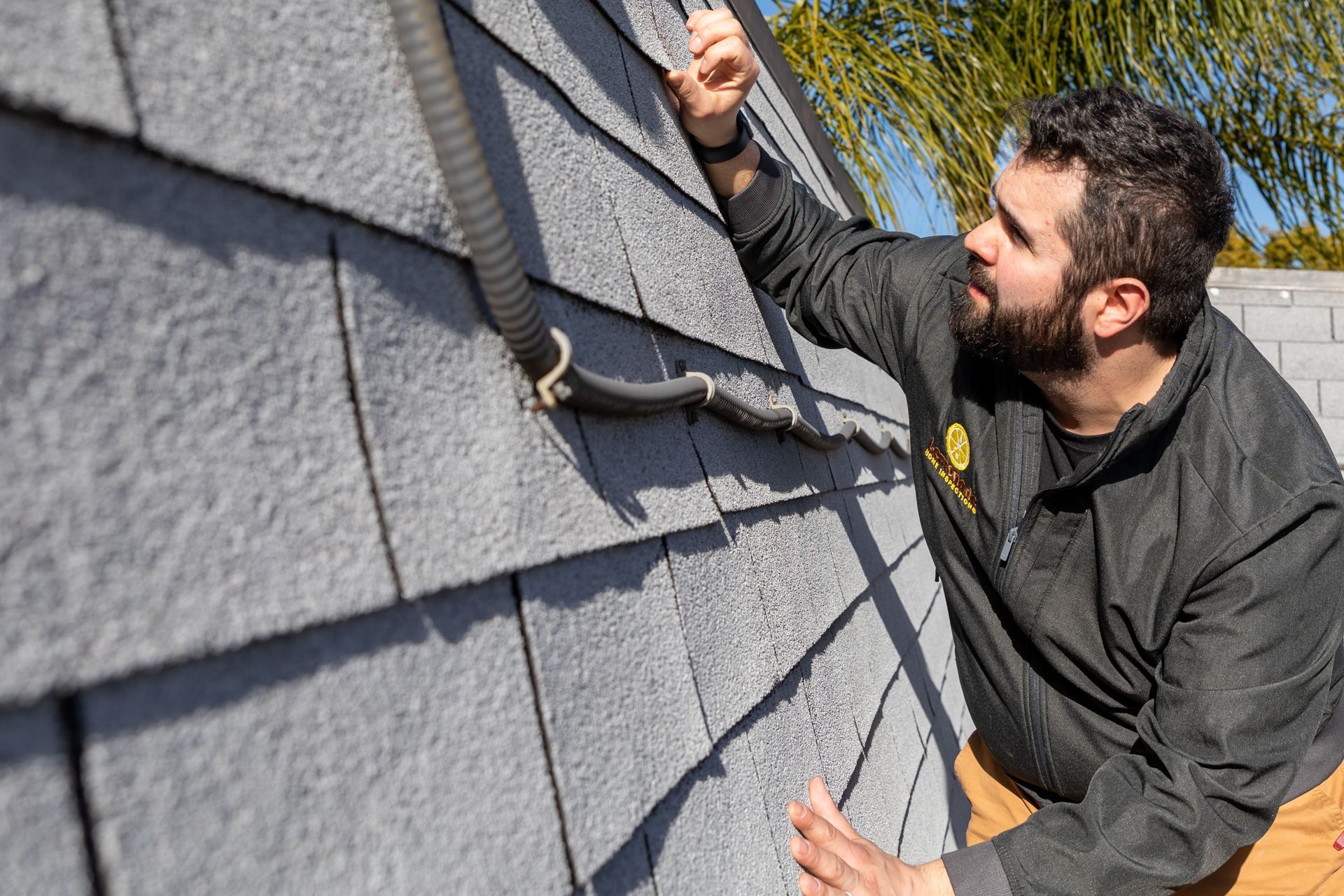 Man inspecting gray roof shingles, holding a black cable. Outdoors in sunlight.