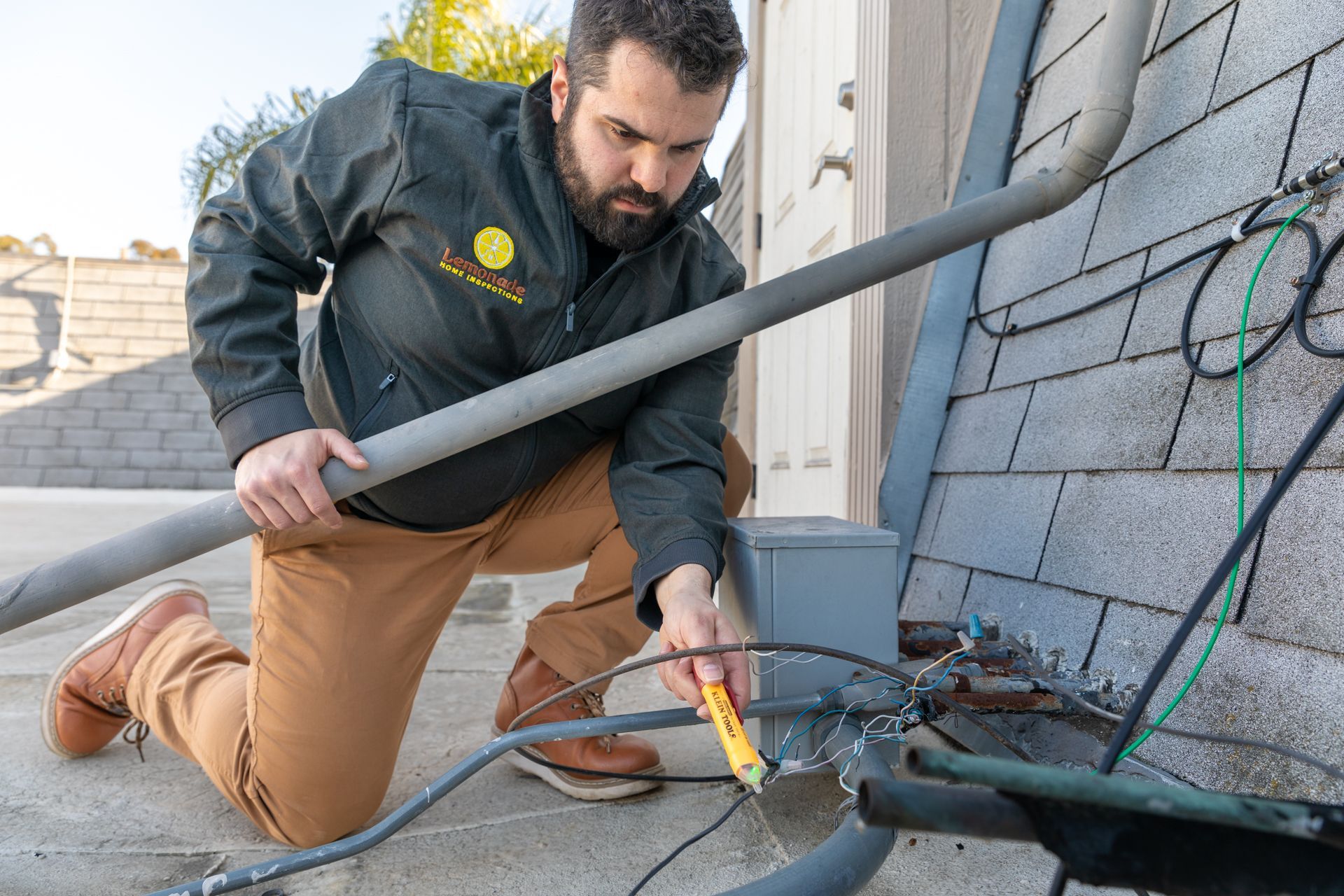 Man in work attire inspecting electrical wiring on a rooftop, using a voltage tester.