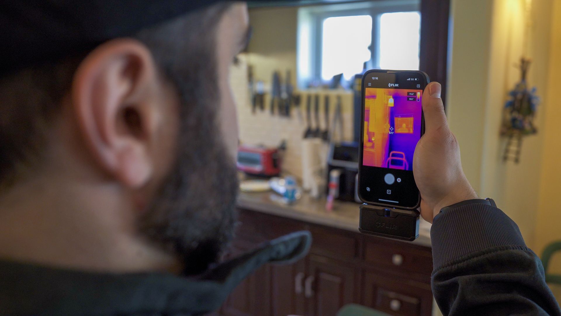 A person holds a smartphone with a thermal imaging attachment in a kitchen, viewing heat patterns on the wall.