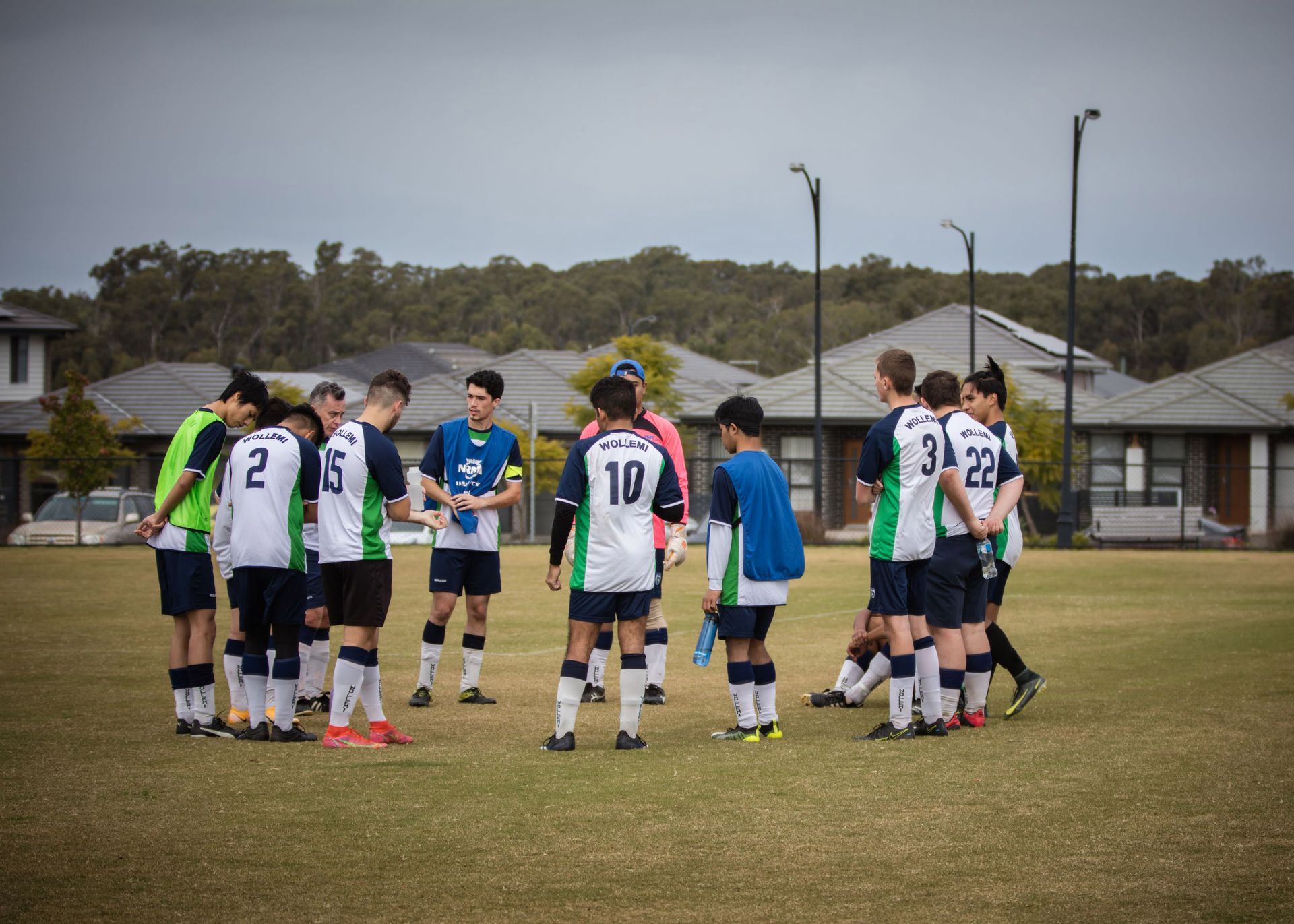 Soccer team huddled on a field, wearing white, green, and blue jerseys; overcast sky, suburban setting.