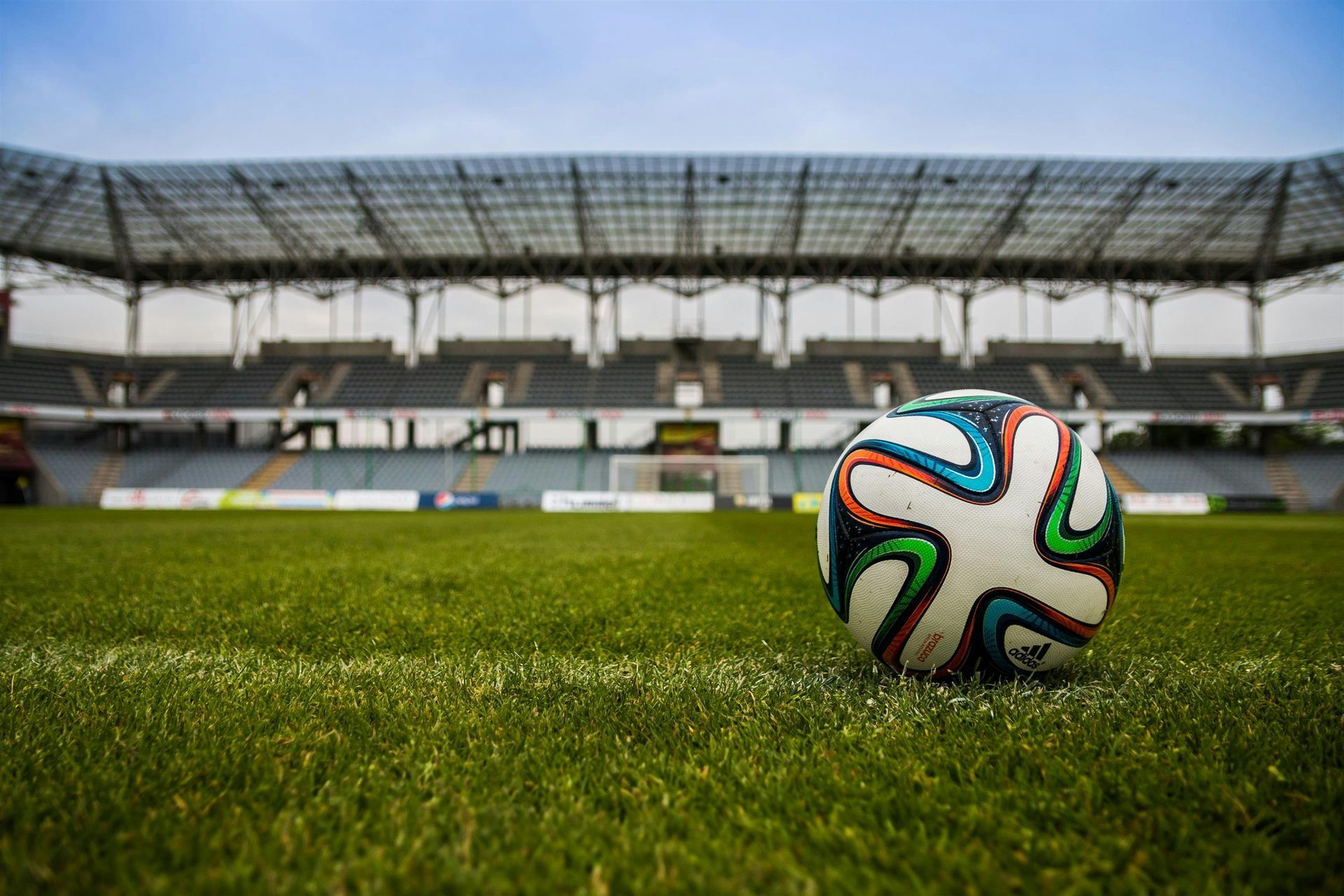 Soccer ball on the field in an empty stadium; green grass, gray seats, and blue sky.