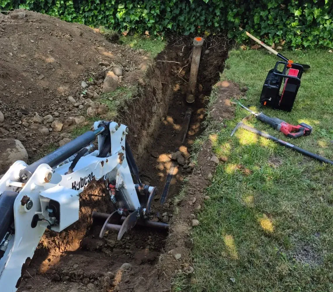 A white excavator digs a trench in a grassy yard, with a saw and other equipment resting on the nearby lawn.
