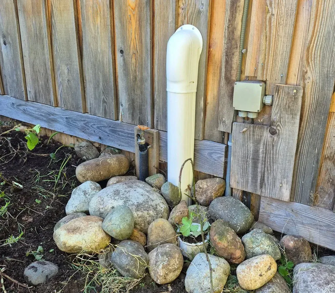 A white PVC pipe stands against a weathered wooden fence, surrounded by a pile of landscaping rocks in a dirt yard.