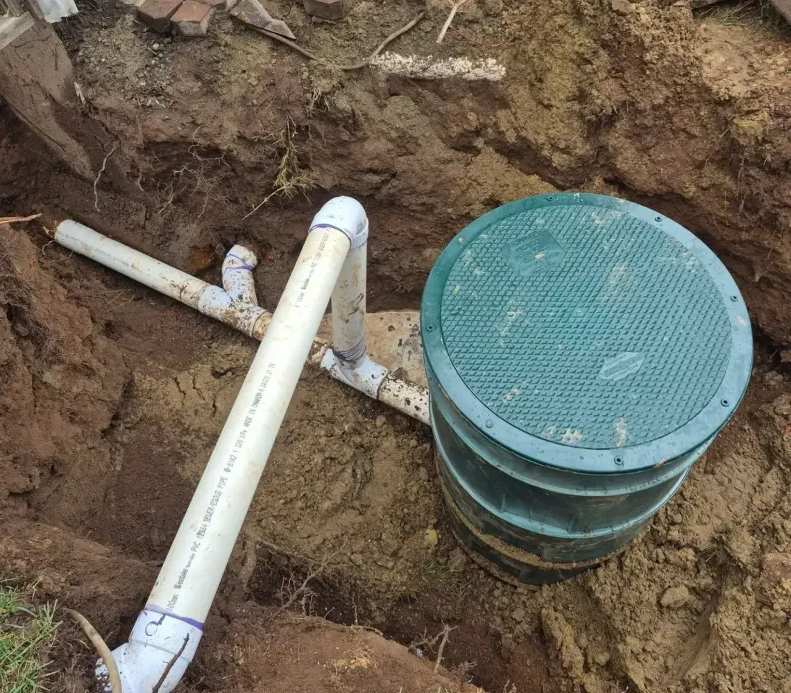 A green-lidded drainage basin installed in an excavated dirt trench, connected to white PVC pipes.