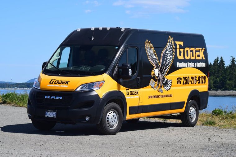 A black and yellow Godek plumbing van with an eagle graphic parked on a gravel shore near water.