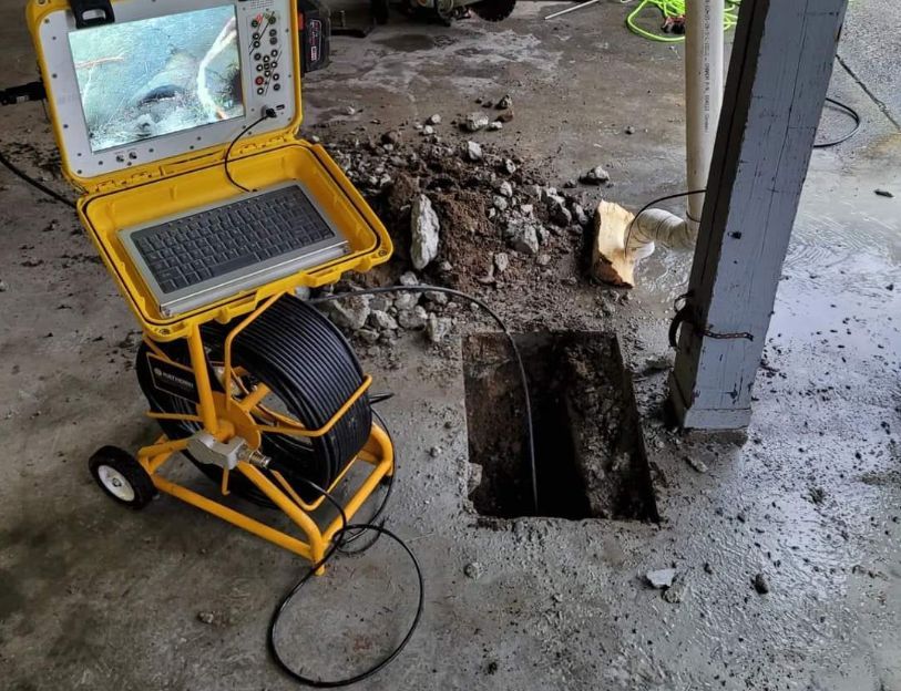 A sewer inspection camera system with a monitor and cable reel positioned next to an open hole in a concrete floor.