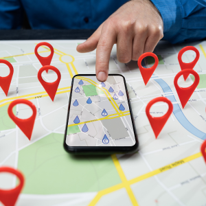A hand points at a smartphone displaying a map with digital pins, placed atop a physical map marked with red location pins.