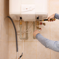 A person repairs the plumbing connections under a white wall-mounted electric water heater using a wrench.