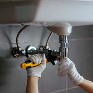 Hands in white work gloves using a tool to tighten the chrome plumbing pipes under a bathroom sink.