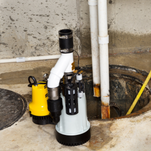 A dual-pump sump pump system with a yellow and a gray motor sitting on a concrete floor next to an open sump pit.