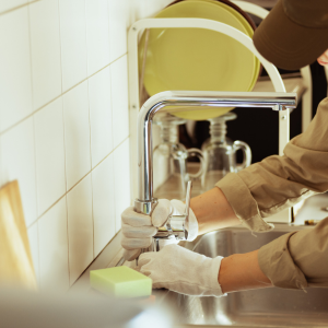 A person wearing white work gloves repairs a kitchen faucet in a sink with white tiled walls and a drying rack nearby.