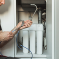 A person installing a water filtration system faucet under a sink, holding the metal spout and connecting blue tubing.