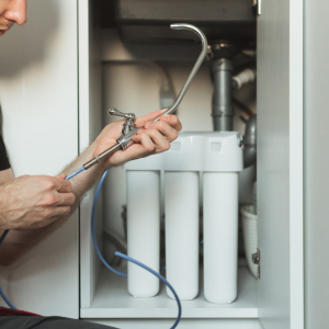 A person installing a water filtration system faucet under a sink, holding the metal spout and connecting blue tubing.