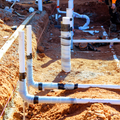 White PVC plumbing pipes emerge from reddish-brown soil in a construction trench for a building foundation.