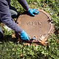 A person in blue nitrile gloves uses a wrench to open a round, heavy-duty iron cover labeled