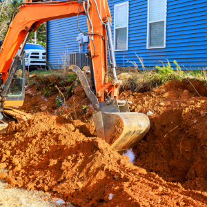 An orange excavator digs a trench in red dirt next to a blue house.