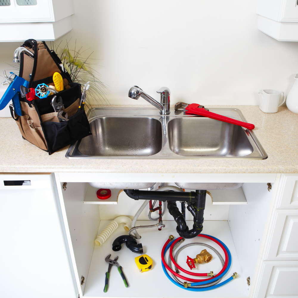 A kitchen sink with a plumbing tool bag on the counter, open cabinets below revealing pipes, tools, and supply lines.