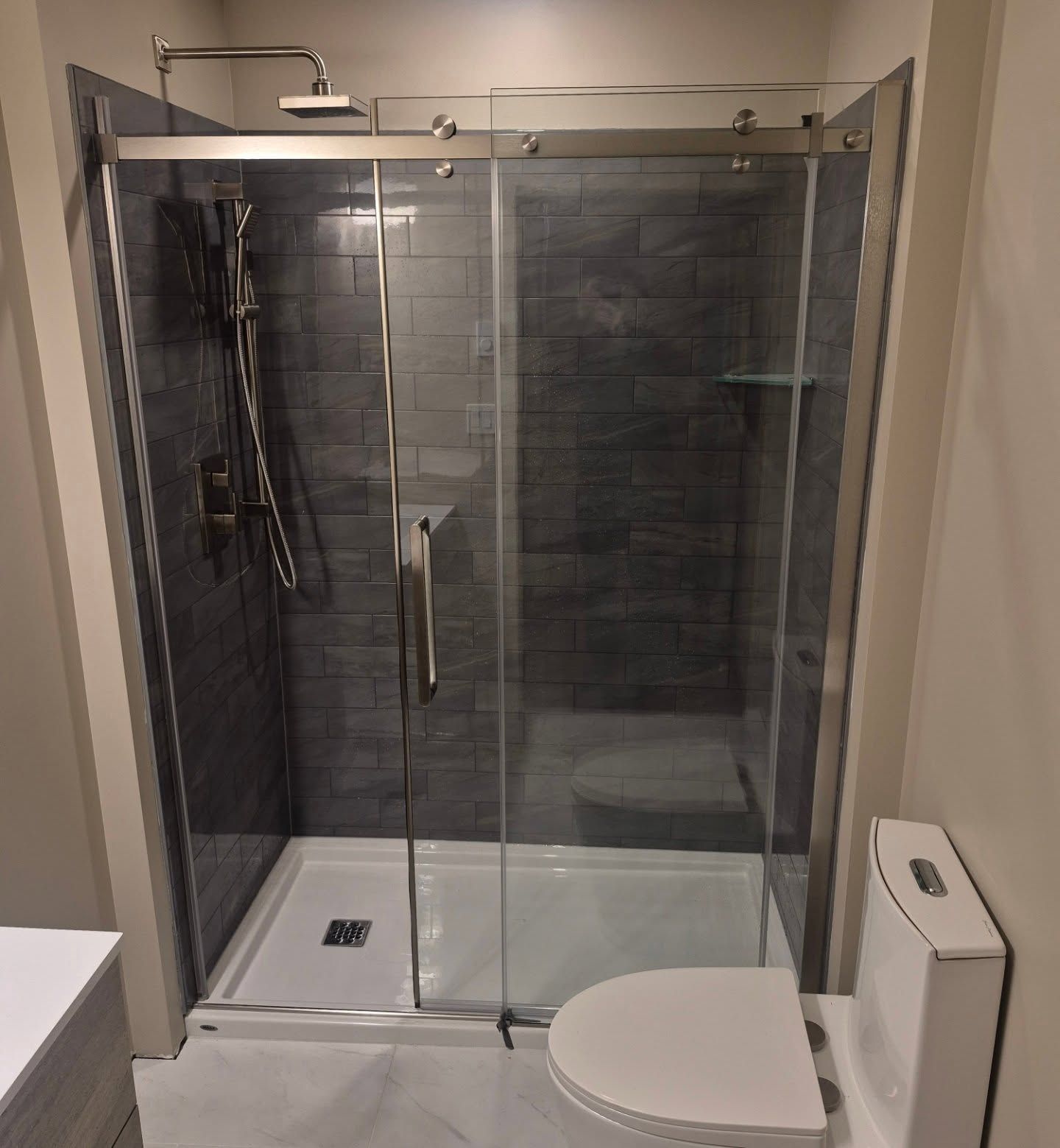 A modern walk-in shower with dark gray subway tiles, a glass sliding door, and a white base, located next to a toilet.