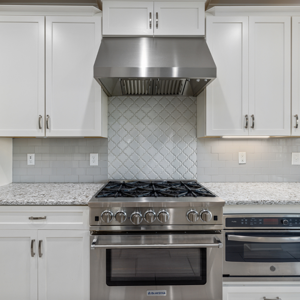 A kitchen stove, oven, and stainless steel range hood set against white cabinetry and a gray tiled backsplash.