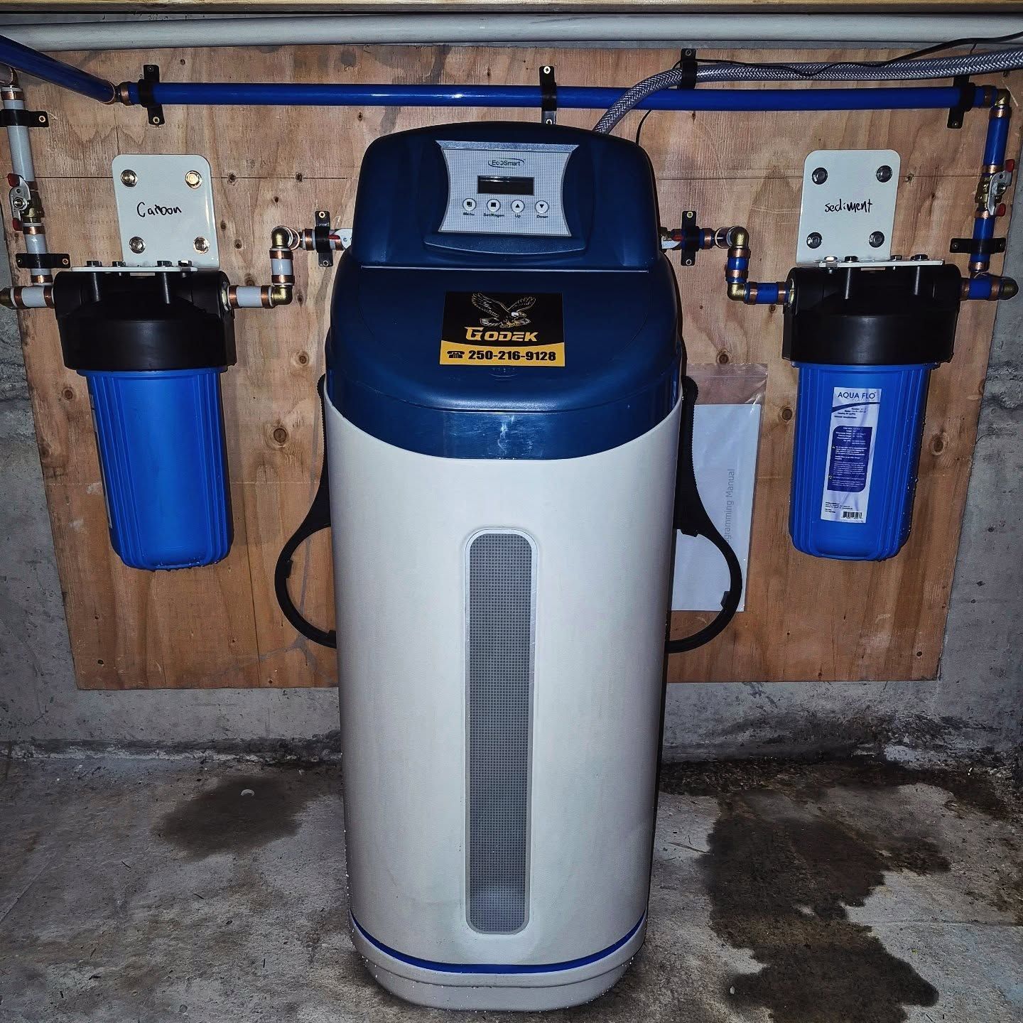 A water filtration system mounted on a plywood wall, featuring two blue canister filters flanking a central softener unit.