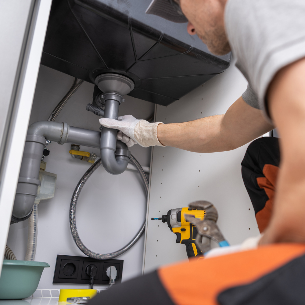 A plumber in work clothes fixing grey plastic sink pipes under a cabinet, with a drill resting nearby.