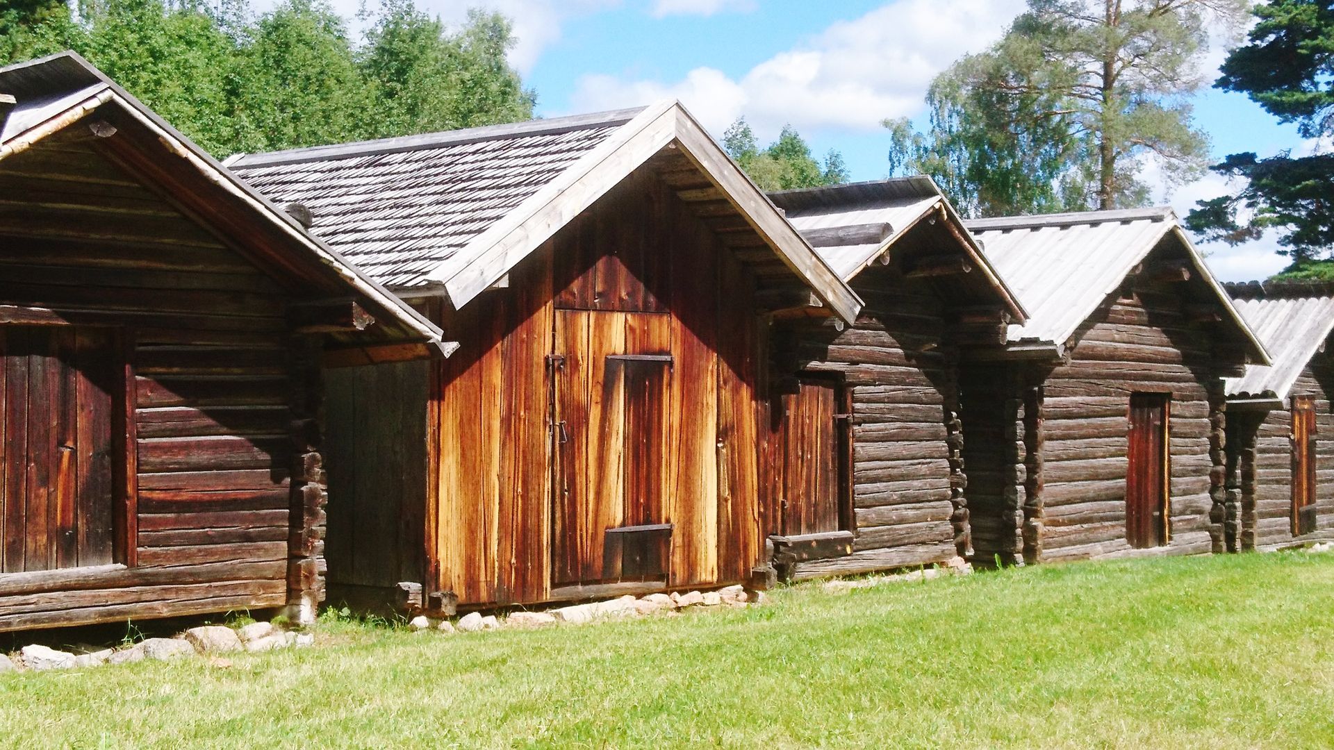 Row of rustic wooden storage sheds with gabled roofs on green grass, surrounded by trees 
