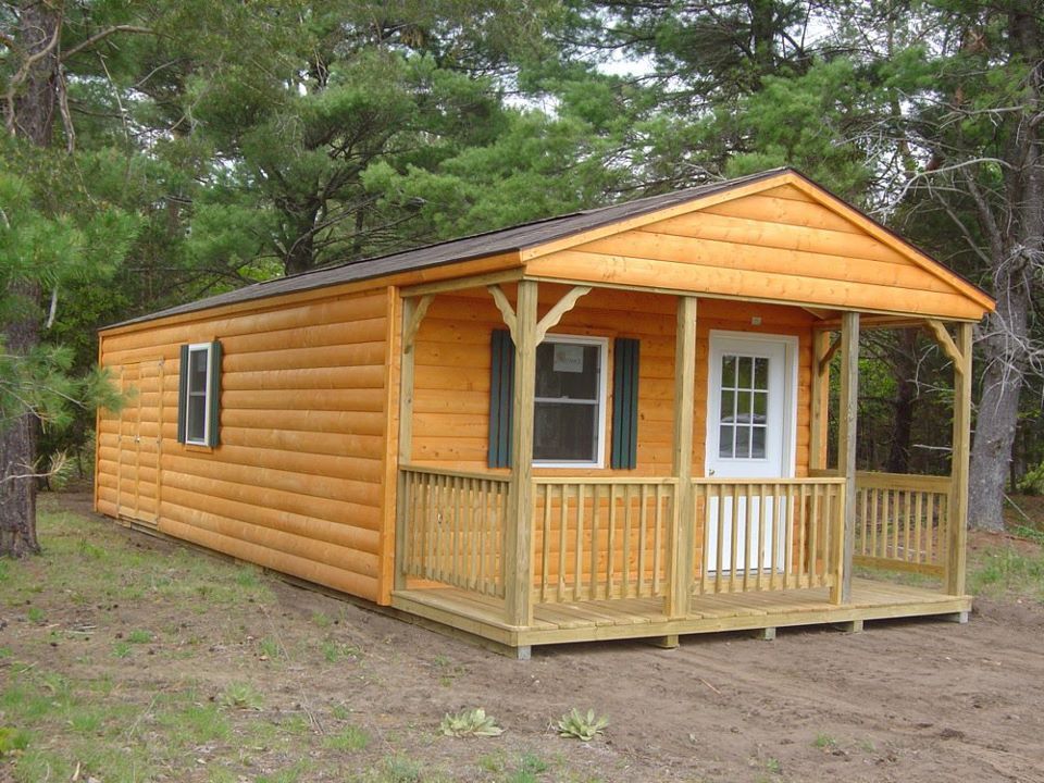 Small wooden cabin with porch, brown siding, white door, and green shutters in a wooded area.