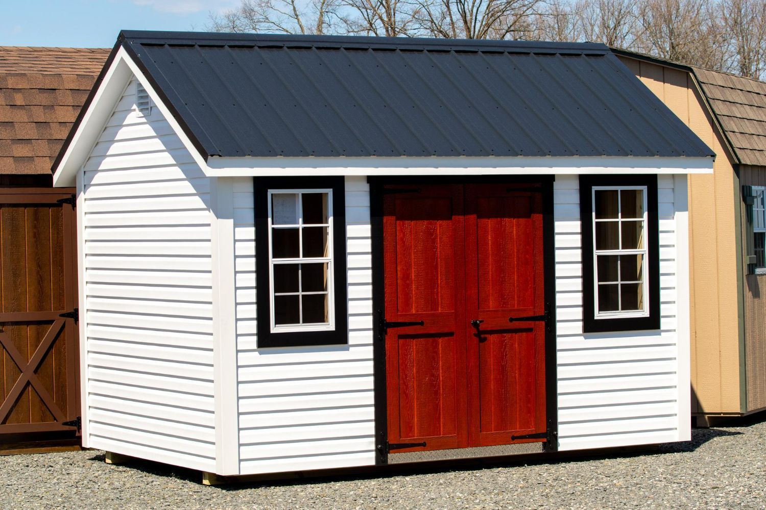White shed with black roof and red double doors, shown outdoors on gravel.