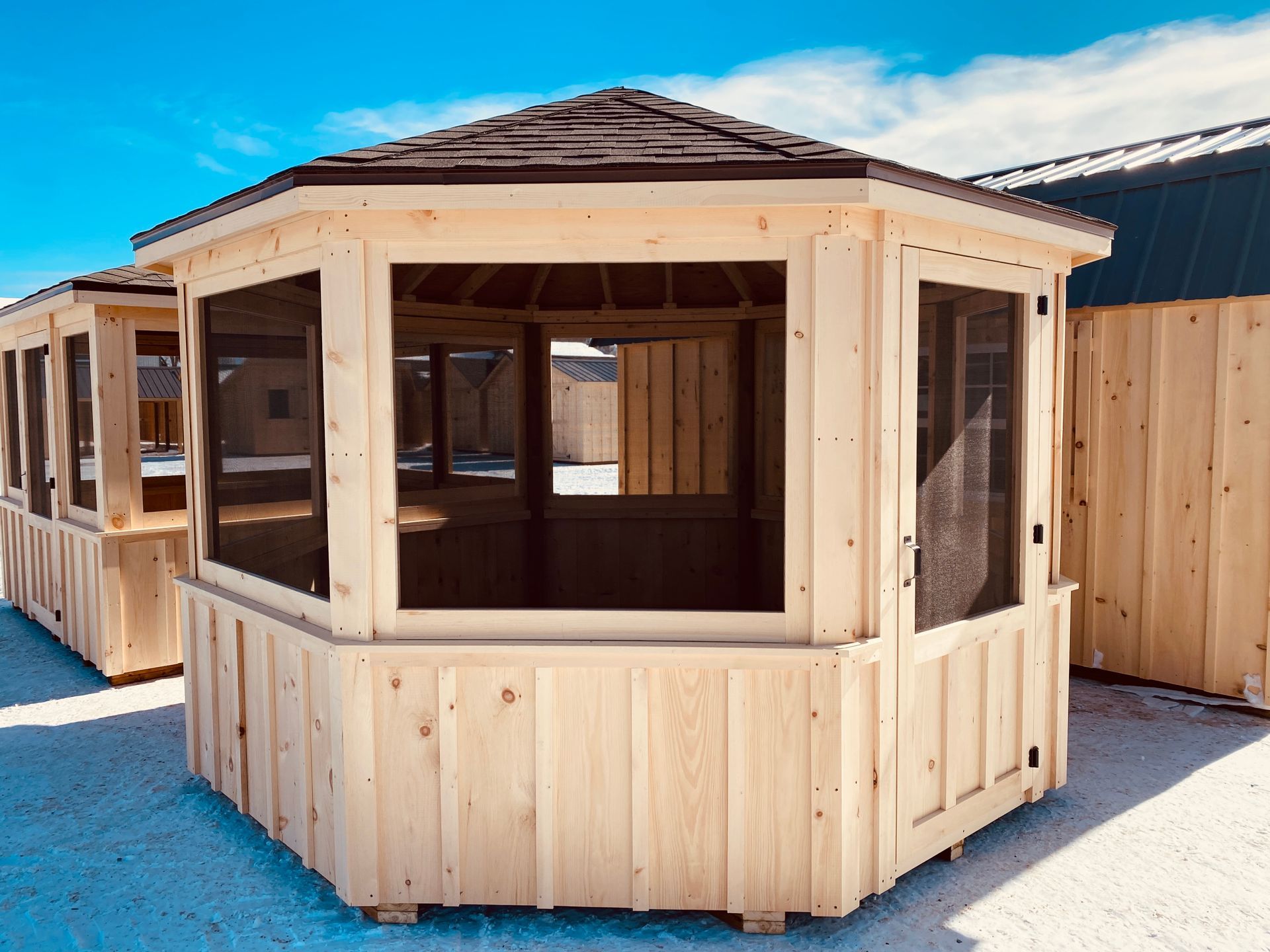 Wooden gazebo with screened windows and a brown shingled roof, set outside on a sunny day.