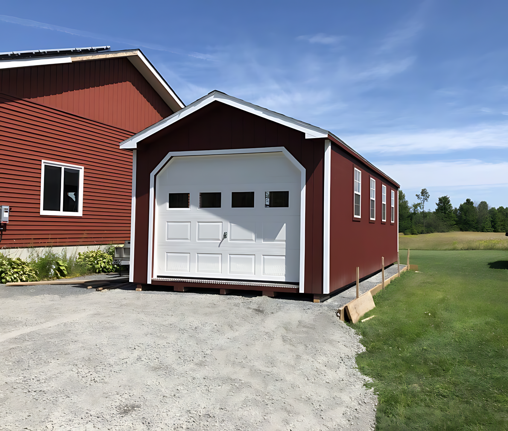 Red garage with white door and trim, next to red building, gravel driveway, grassy field.