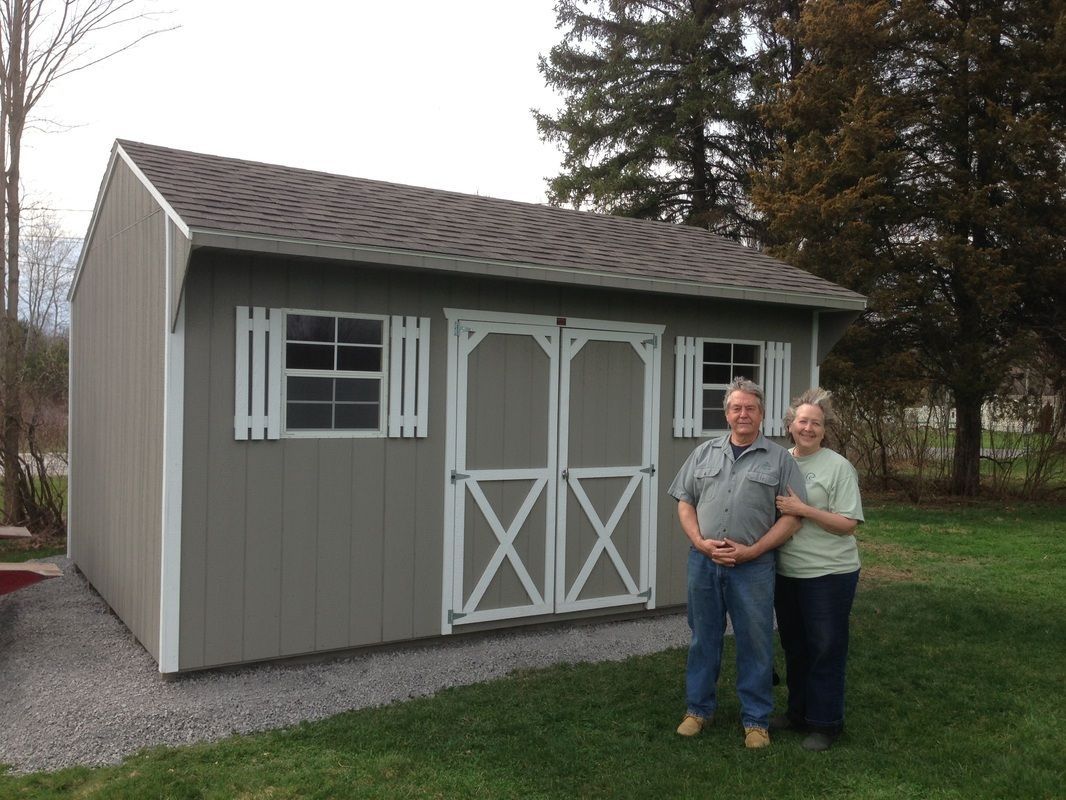 Couple stands in front of a new gray shed with white trim and a brown roof, on a gravel base in a grassy yard.