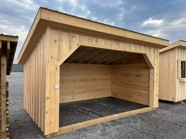 Wooden open-front shed with light-colored siding, a brown roof, and a gravel floor, under a cloudy sky.