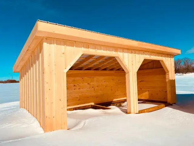 Wooden, open-sided shelter in snowy field, under a bright blue sky.