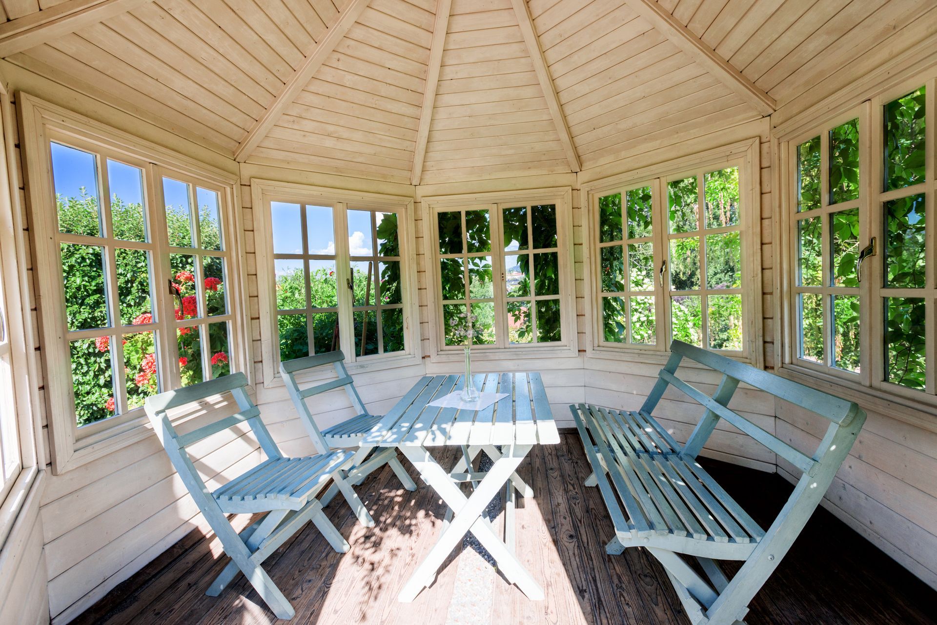 White gazebo interior with light blue furniture, surrounded by windows with a garden view.