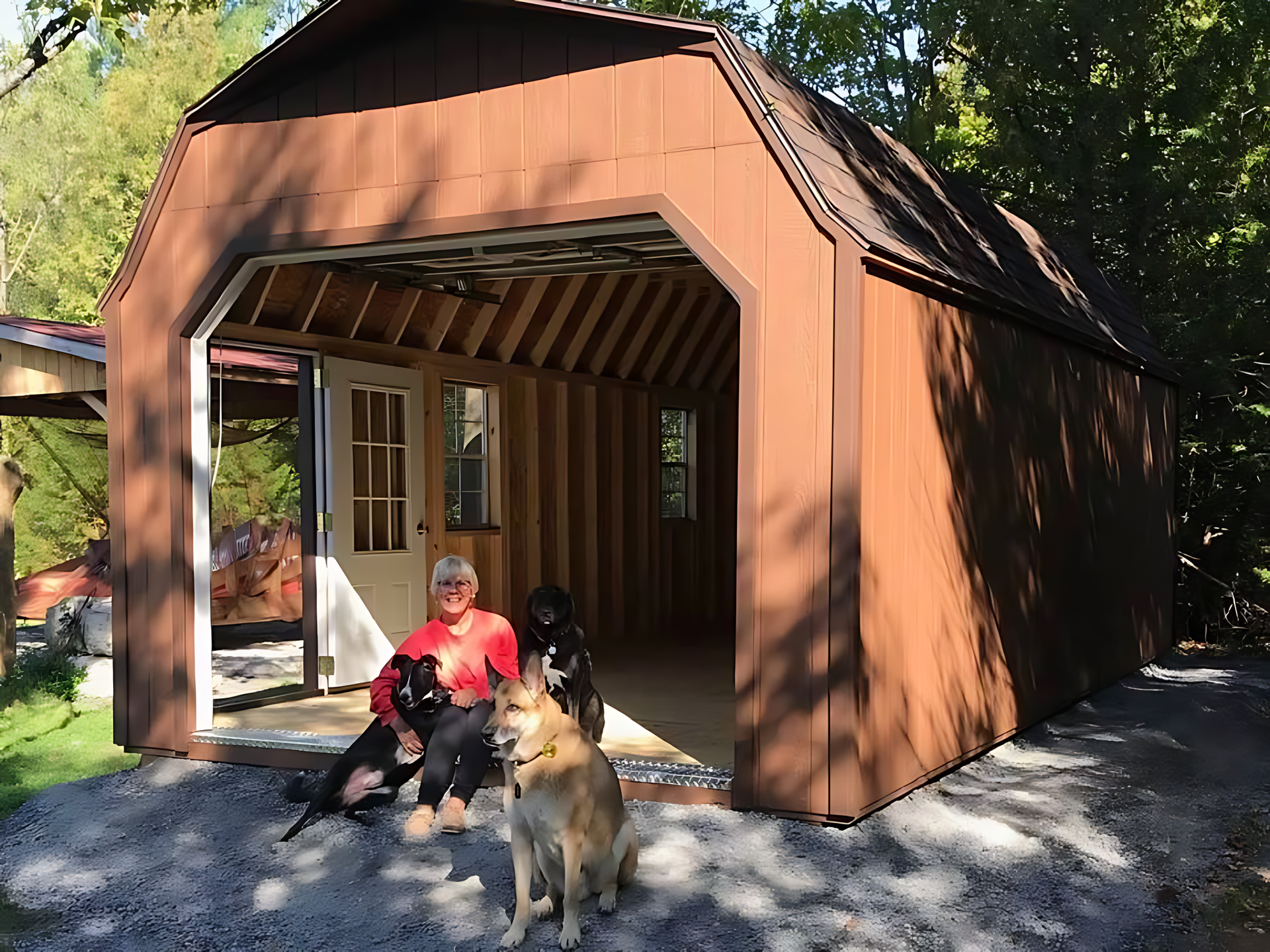 A woman sits in front of a brown garage with two dogs