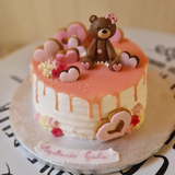 Torta con un orsacchiotto sopra, glassa rosa, decorazioni a forma di cuore.