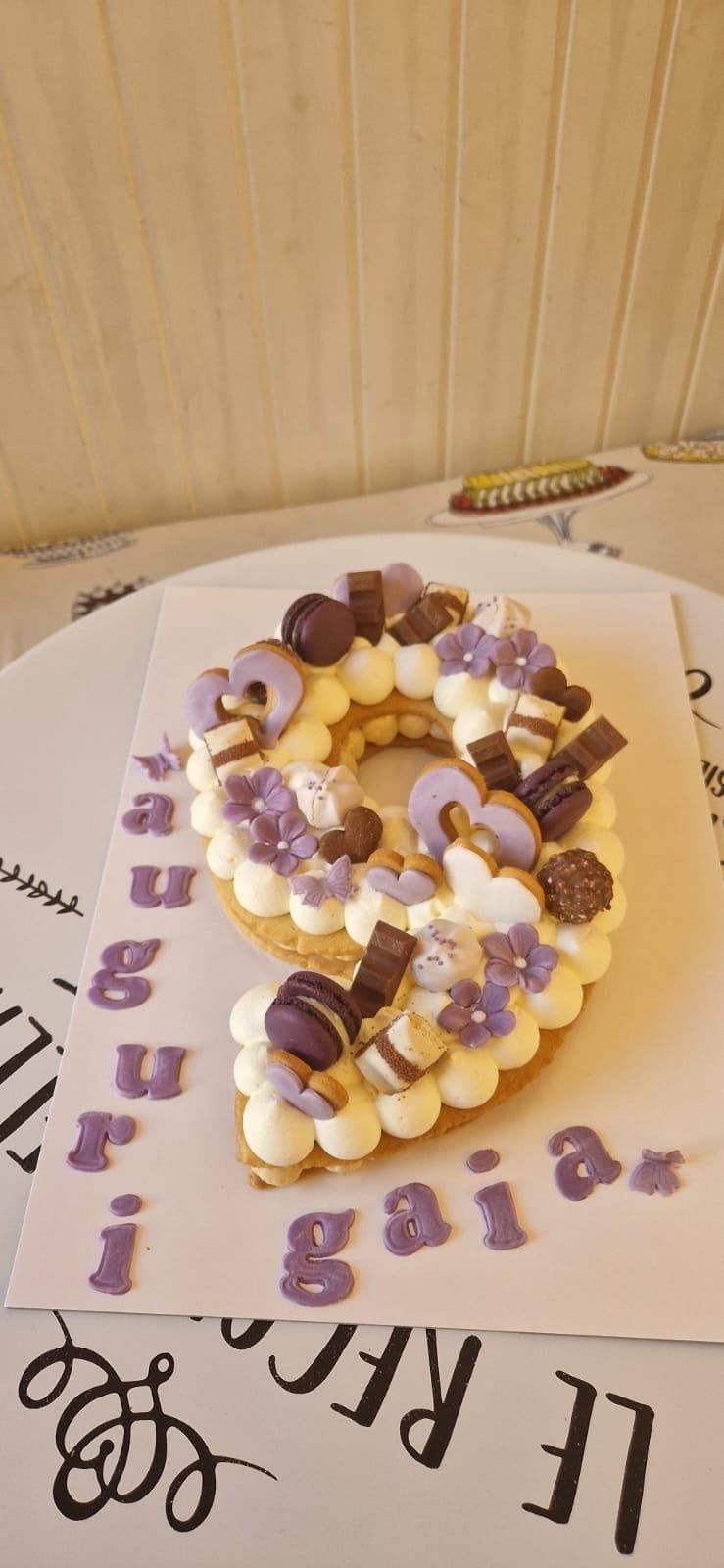 Torta a forma di numero 9, decorata con fiori viola, macarons e cioccolato.