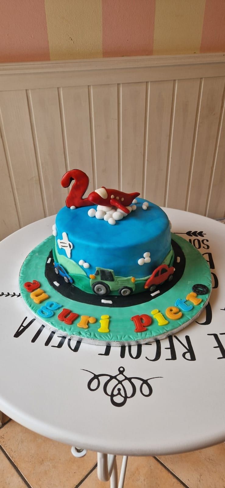 Torta decorata con un cielo azzurro, un aeroplano rosso e veicoli con la scritta