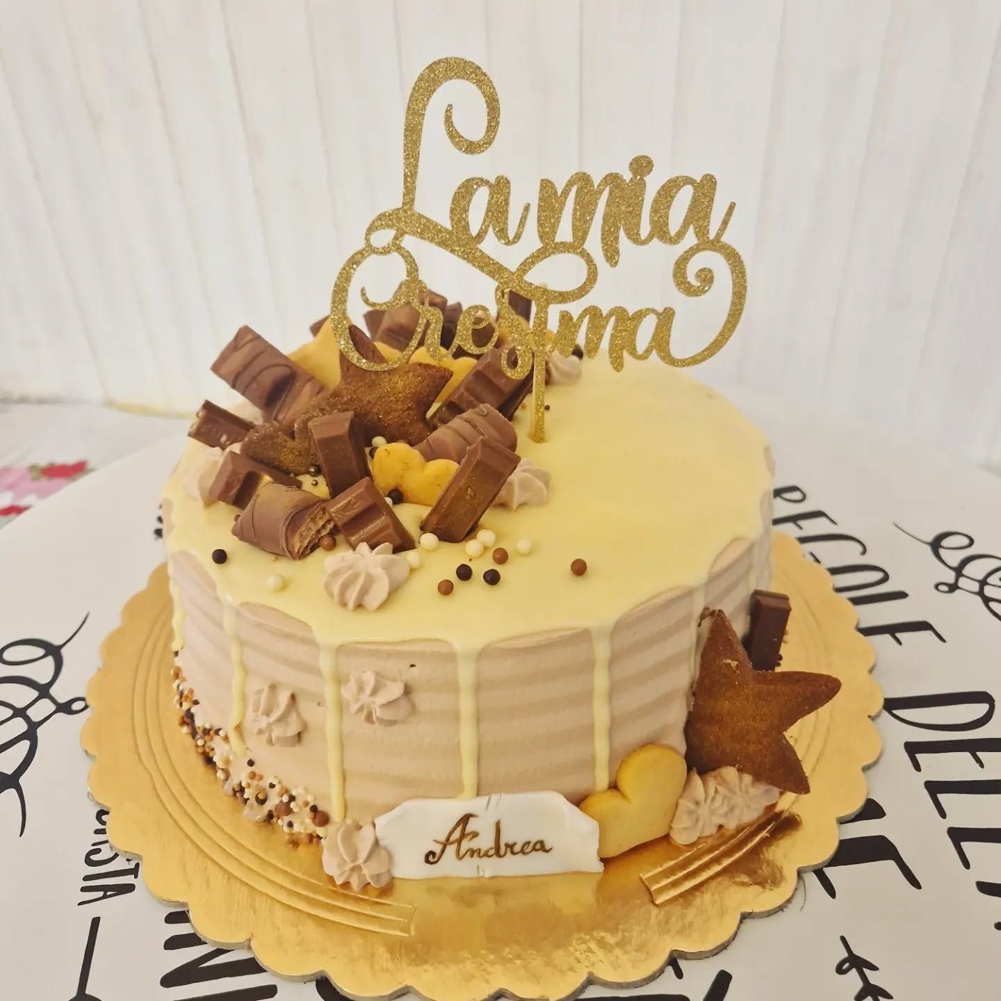 Torta decorata con pezzetti di cioccolato, biscotti a forma di stella e a forma di cuore e un topper dorato con la scritta