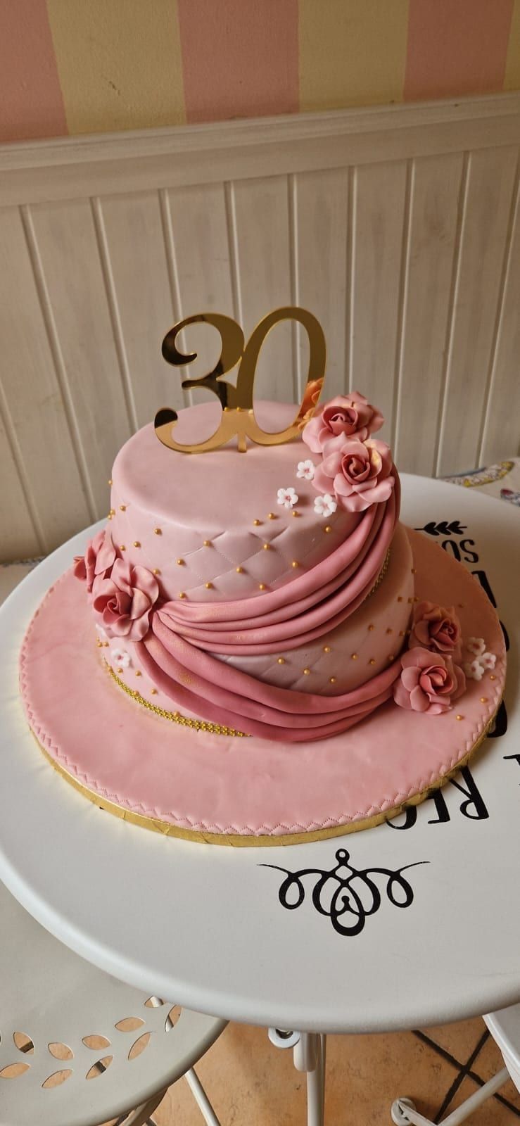 Torta a più piani rosa con dettagli floreali e un topper dorato con la scritta
